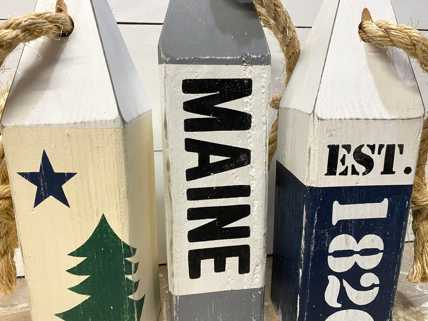 Close-up of decorative Maine nautical buoy showing hand-painted original Maine flag details, established date, distressed wood finish, and rustic coastal decor style