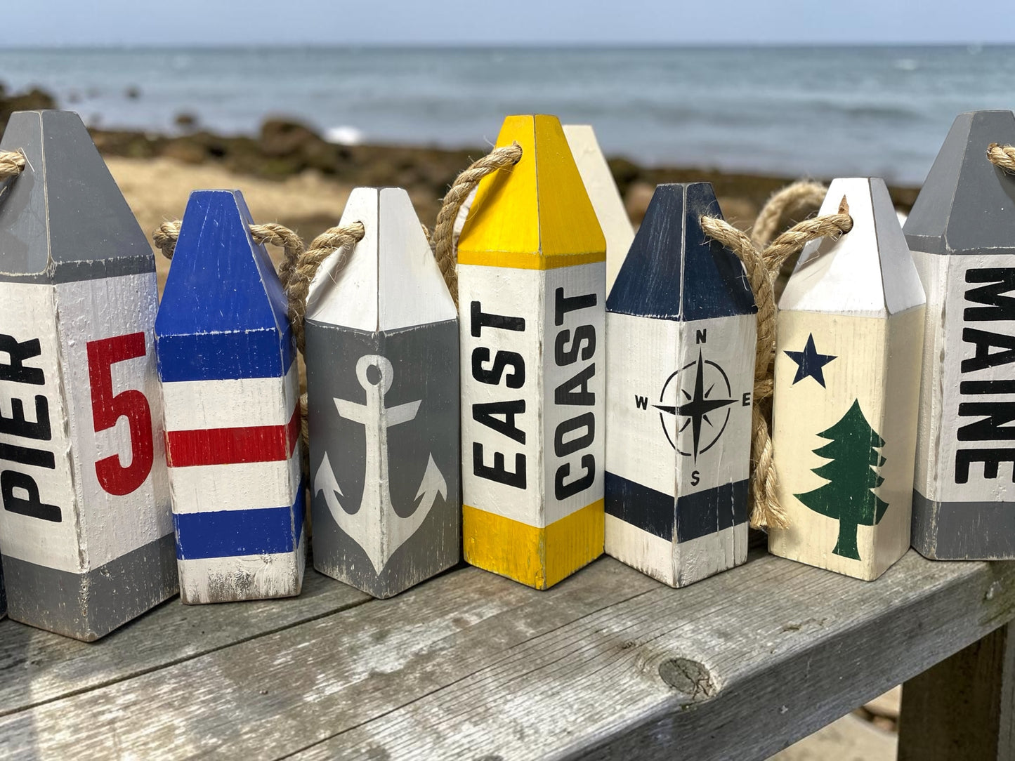 Decorative East Coast wood buoy set displayed near the ocean featuring compass rose and anchor designs. Coastal decor inspired by seaside living, beach houses, cottages, and waterfront spaces
