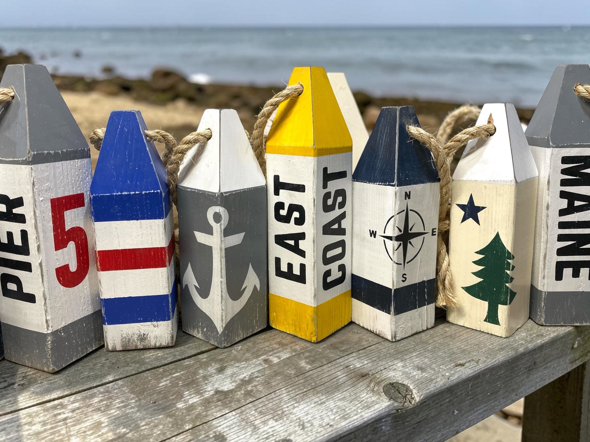 Decorative East Coast wood buoy set displayed near the ocean featuring compass rose and anchor designs. Coastal decor inspired by seaside living, beach houses, cottages, and waterfront spaces
