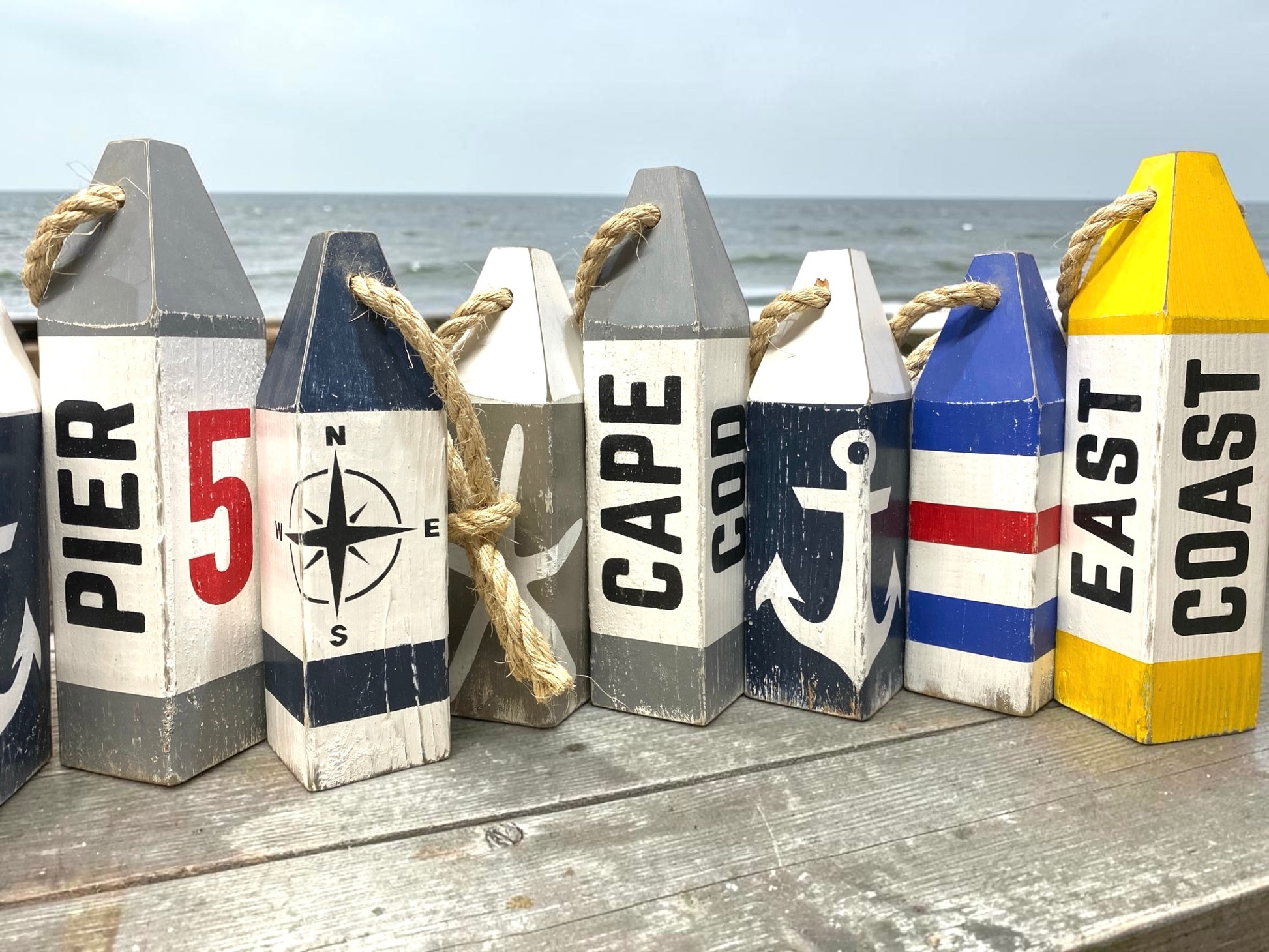 Cape Cod wood buoy set displayed near the ocean featuring hand-painted starfish and anchor designs with a rustic distressed finish. Nautical coastal decor for beach houses, coastal cottages, beach-themed rooms, seaside homes, and waterfront spaces.