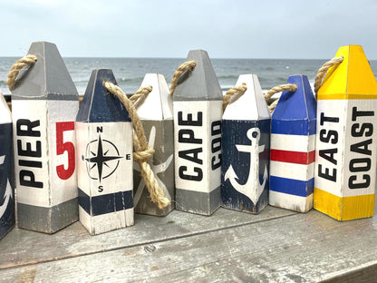 Cape Cod wood buoy set displayed near the ocean featuring hand-painted starfish and anchor designs with a rustic distressed finish. Nautical coastal decor for beach houses, coastal cottages, beach-themed rooms, seaside homes, and waterfront spaces.