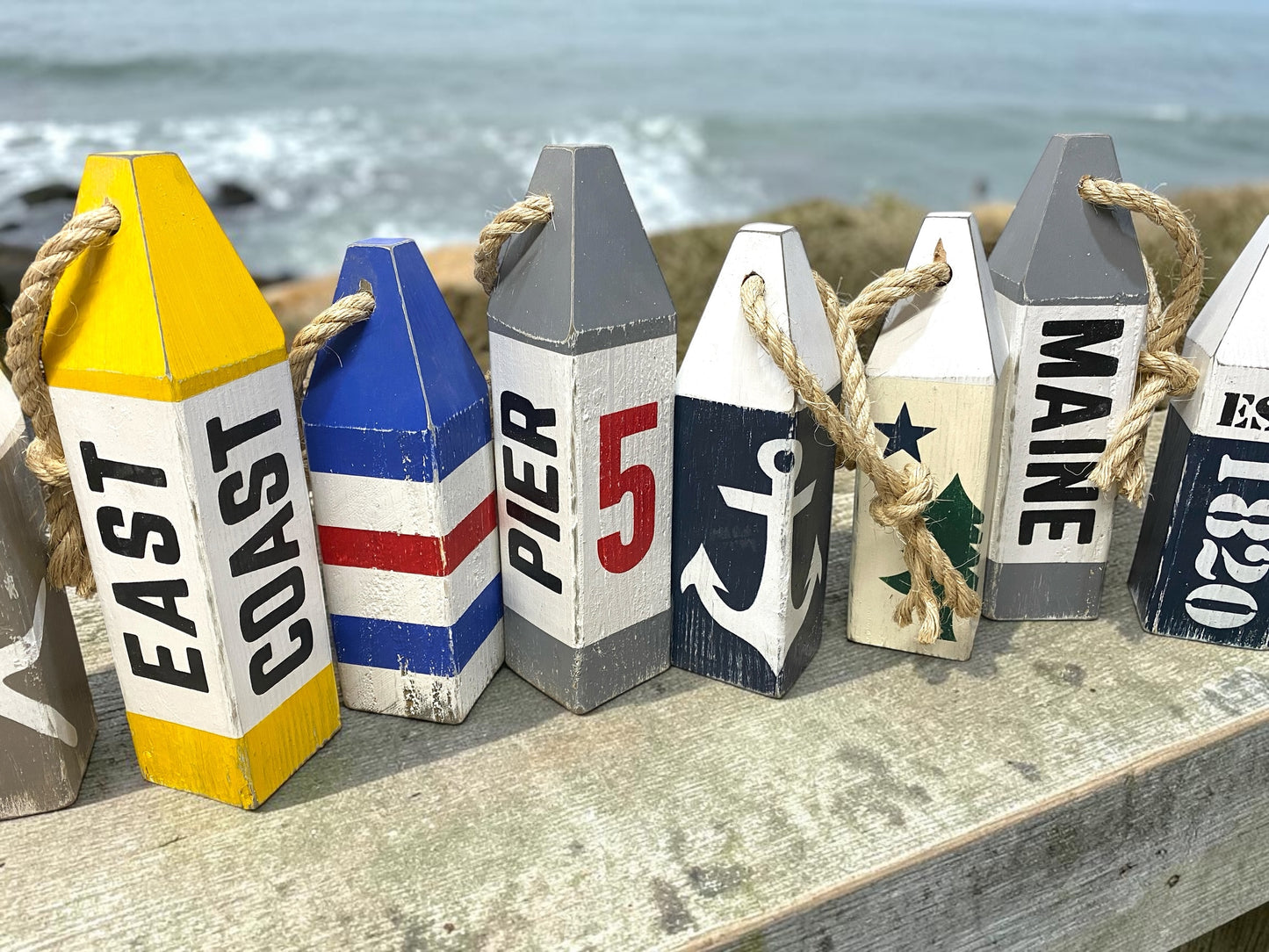 Hand-painted Pier 5 lobster wood buoys featuring nautical coastal designs for beach house decor, lake homes, cottages, and boating enthusiasts. Shown at the ocean with other buoy sets