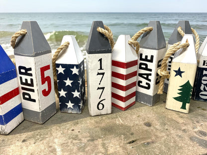 Decorative 1776 American flag wood buoy set displayed near the ocean with patriotic coastal style. Nautical decor inspired by seaside living, beach houses, harbors, and Americana design